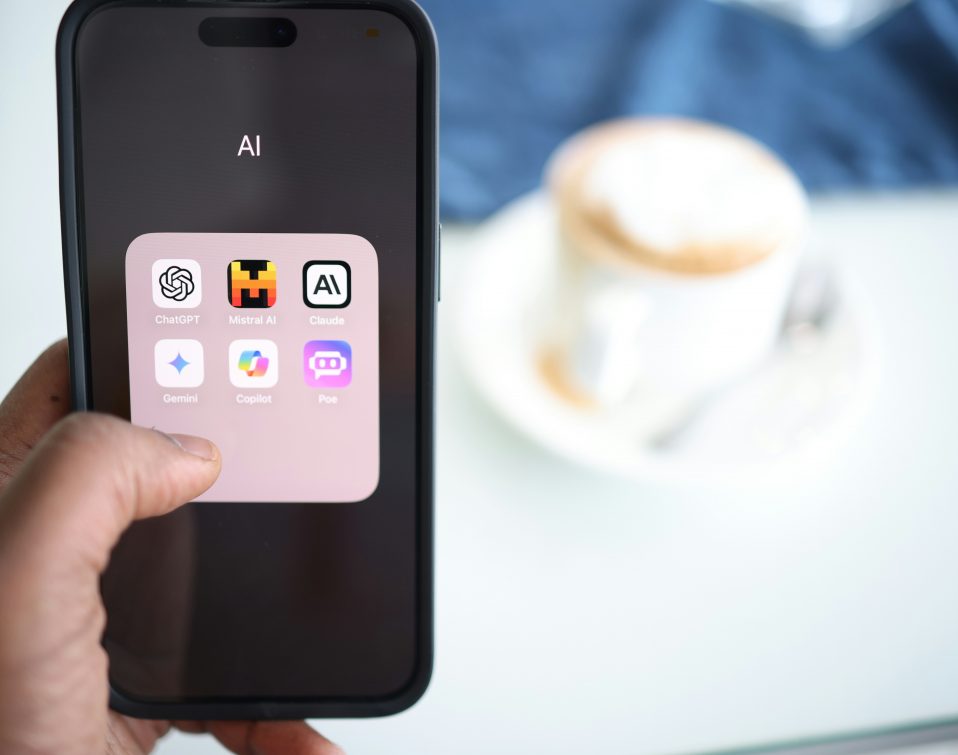 Hand holding a smartphone displaying a folder of AI apps including ChatGPT, Mistral AI, Claude, Gemini, Copilot, and Poe