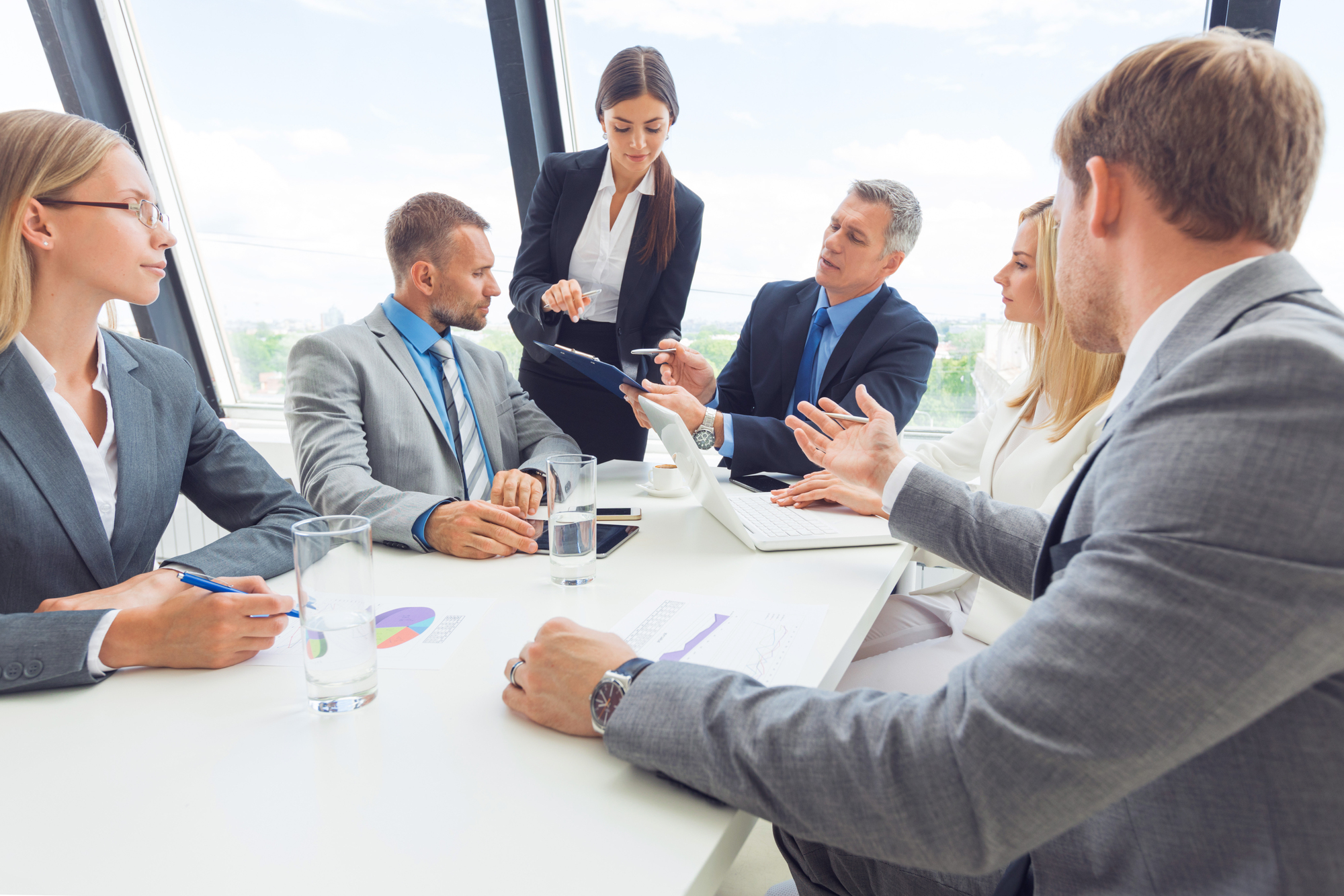 Multi-stakeholder business meeting with five professionals reviewing documents and discussing a decision around a conference table
