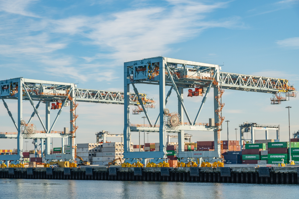 A real photo of a busy shipping port with large gantry cranes and stacked intermodal shipping containers &mdash; including visible Evergreen and CMA CGM branding. Water in the foreground, blue sky with light clouds. Clean, professional shot.